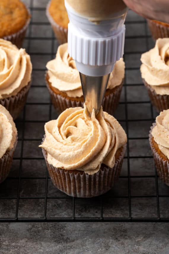 A piping tip pipes a swirl of frosting over a banana cupcake on a wire rack, surrounded by more frosted cupcakes.