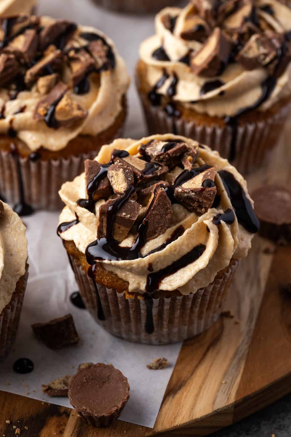 Close up of banana cupcakes topped with frosting and a drizzle of chocolate on a parchment-lined wooden board.