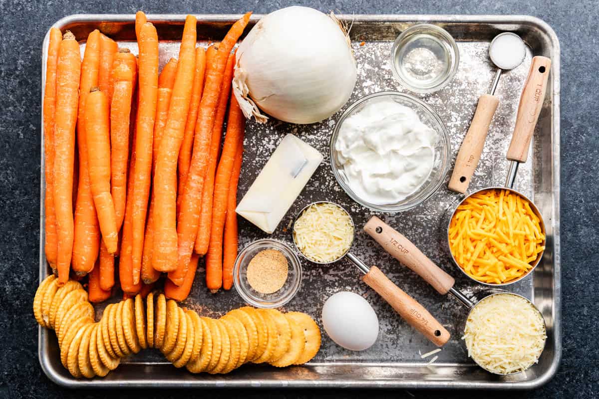 An overhead view of a sheet pan holding raw, wet and dry ingredients needed to make cheesy carrot casserole.