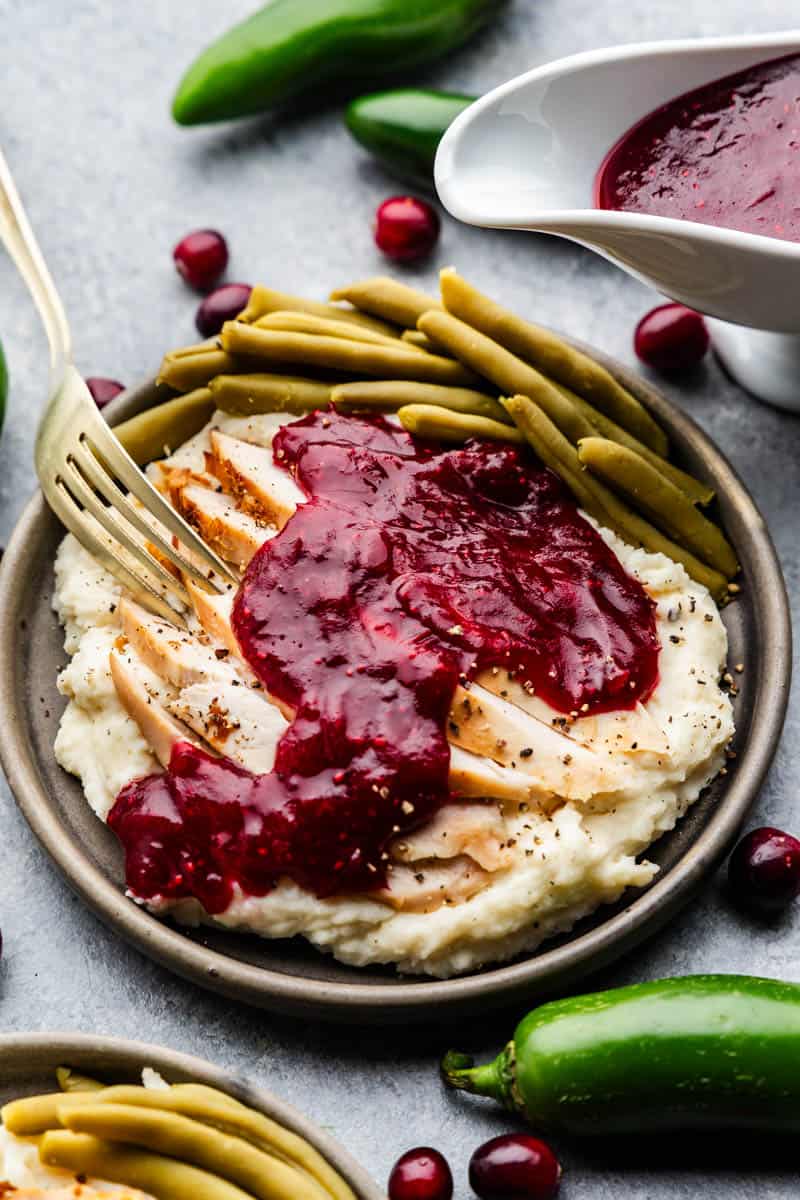 A plate filled with mashed potatoes topped with slices of turkey, covered with jalapeno cranberry sauce, next to green beans, with a fork set on the side of the plate.