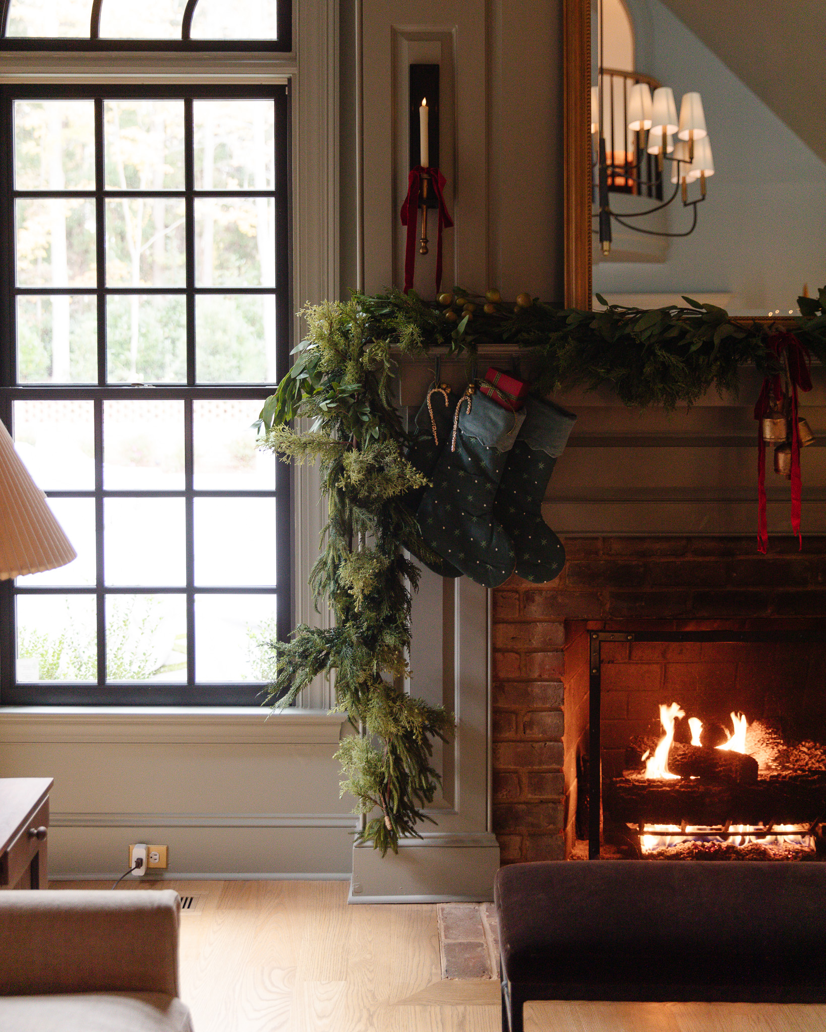 An oversized garland of greenery on a mantle over a roaring fire behind pine green stockings.