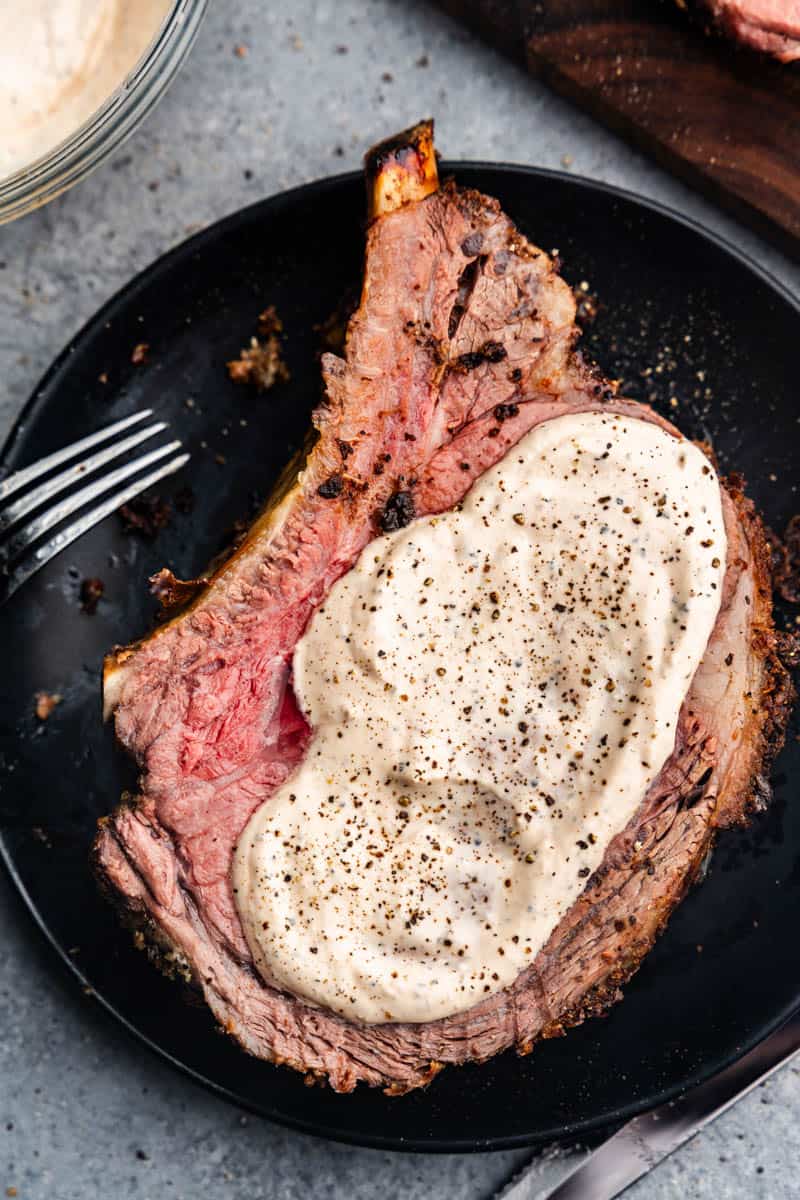 A black plate with a slice of medium-rare prime rib smothered in a horseradish sauce.