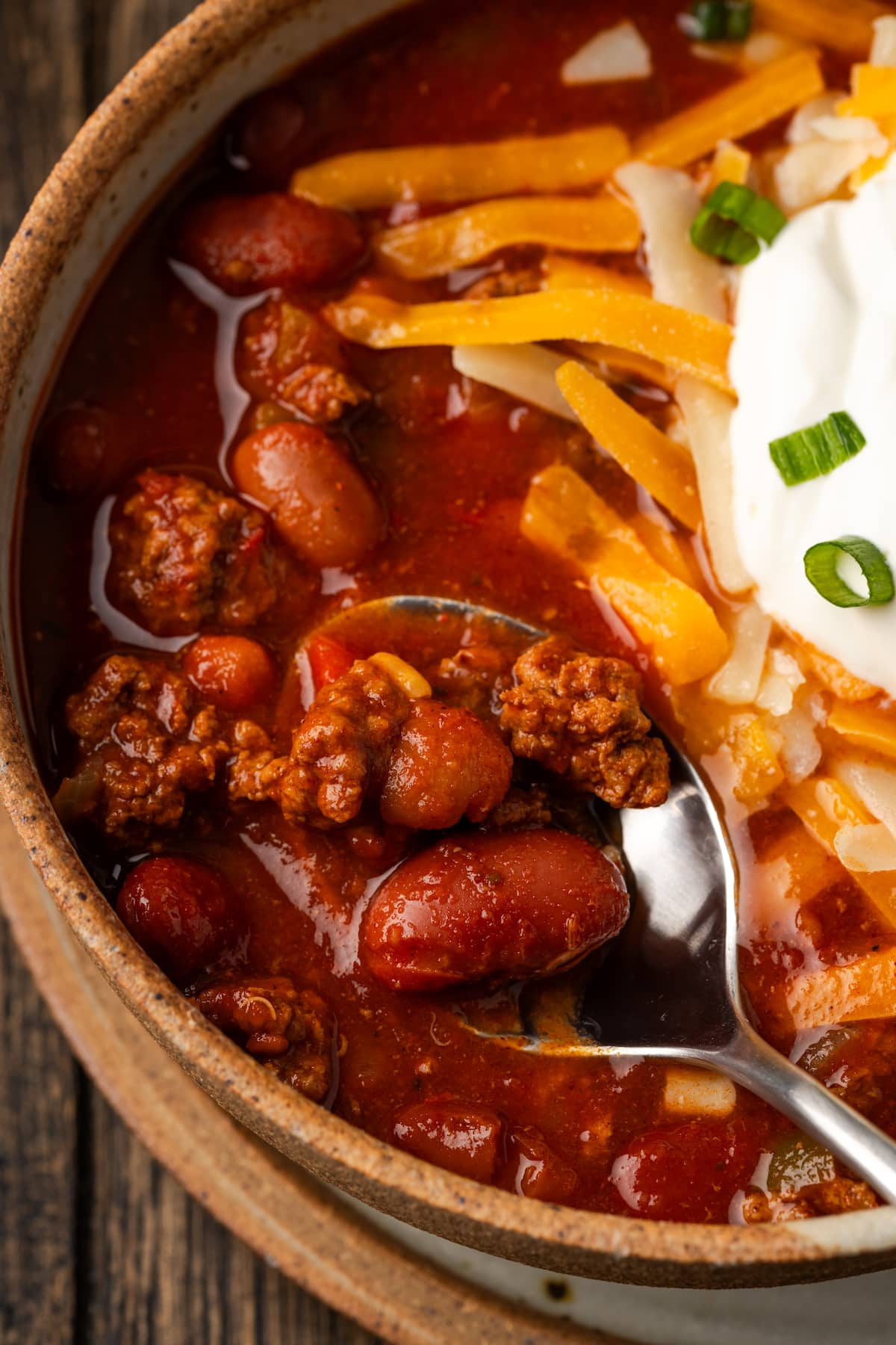 Close up of a spoonful of chili held over a bowlful garnished with shredded cheese, sour cream, and chives.