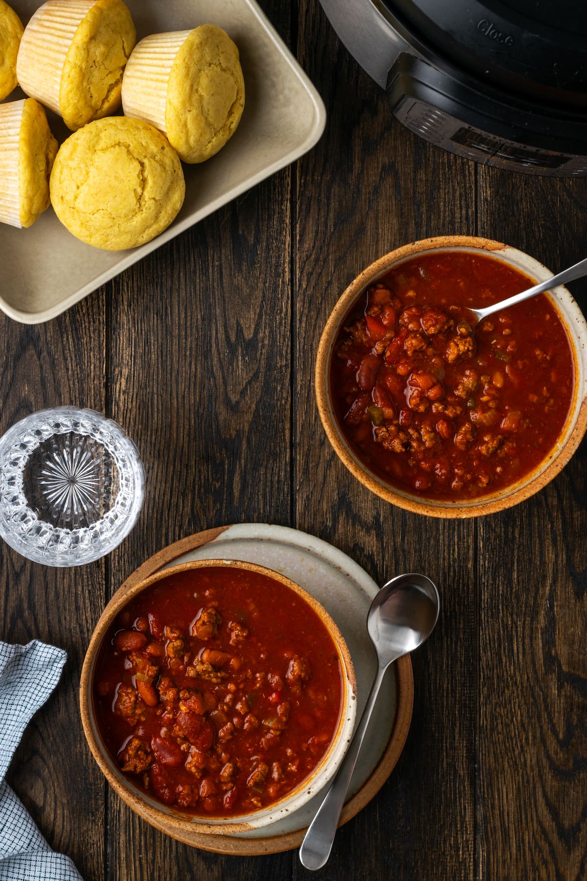 Overhead view of two bowls of chili next to a plate of cornbread muffins on a wooden countertop.