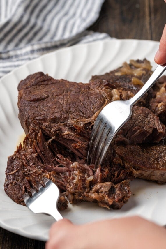 Two forks are used to shred one corner of an instant pot chuck roast on a plate.