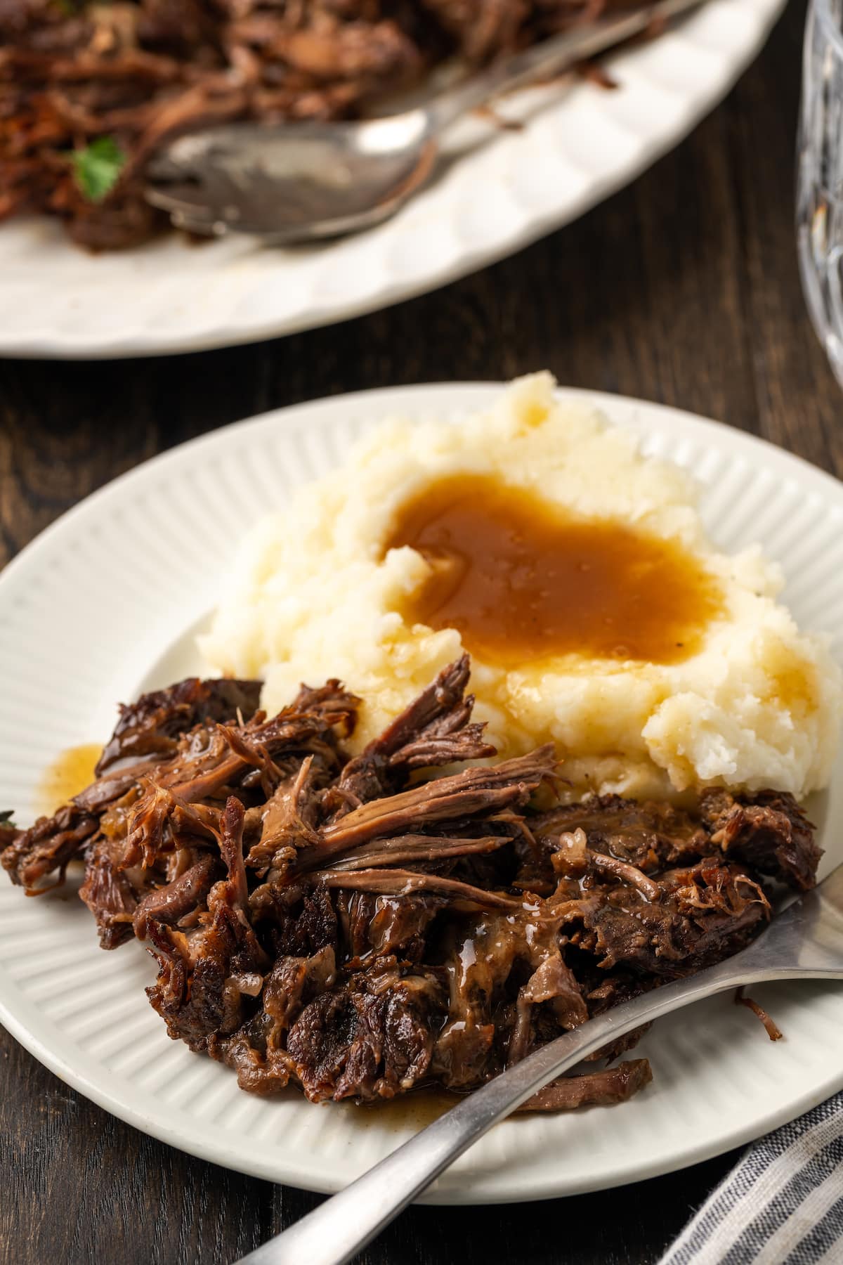 Shredded pot roast served on a white plate next to mashed potatoes covered in gravy, with more shredded roast on a plate in the background.