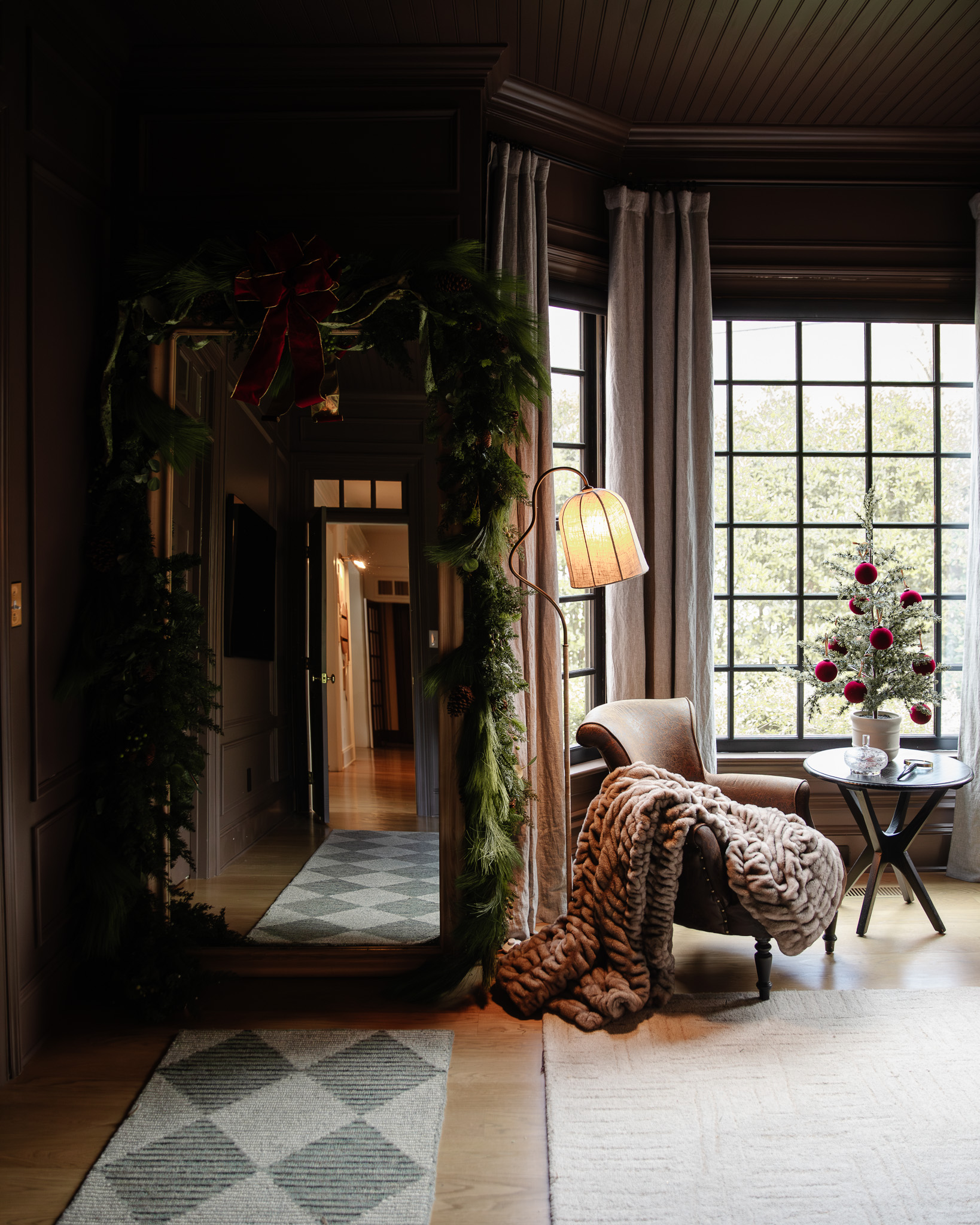 The sitting nook of a primary bedroom decorated for Christmas with a tabletop tree, greenery, and a plush brown blanket over the leather chair