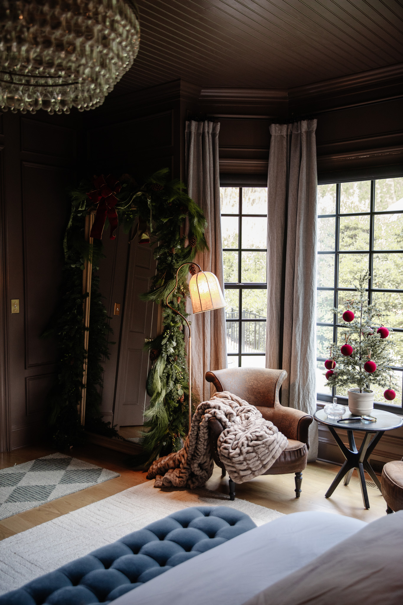 The sitting nook of a primary bedroom decorated for Christmas with a tabletop tree and red velvet baubles, and a large greenery swag over the oversized leaning mirror