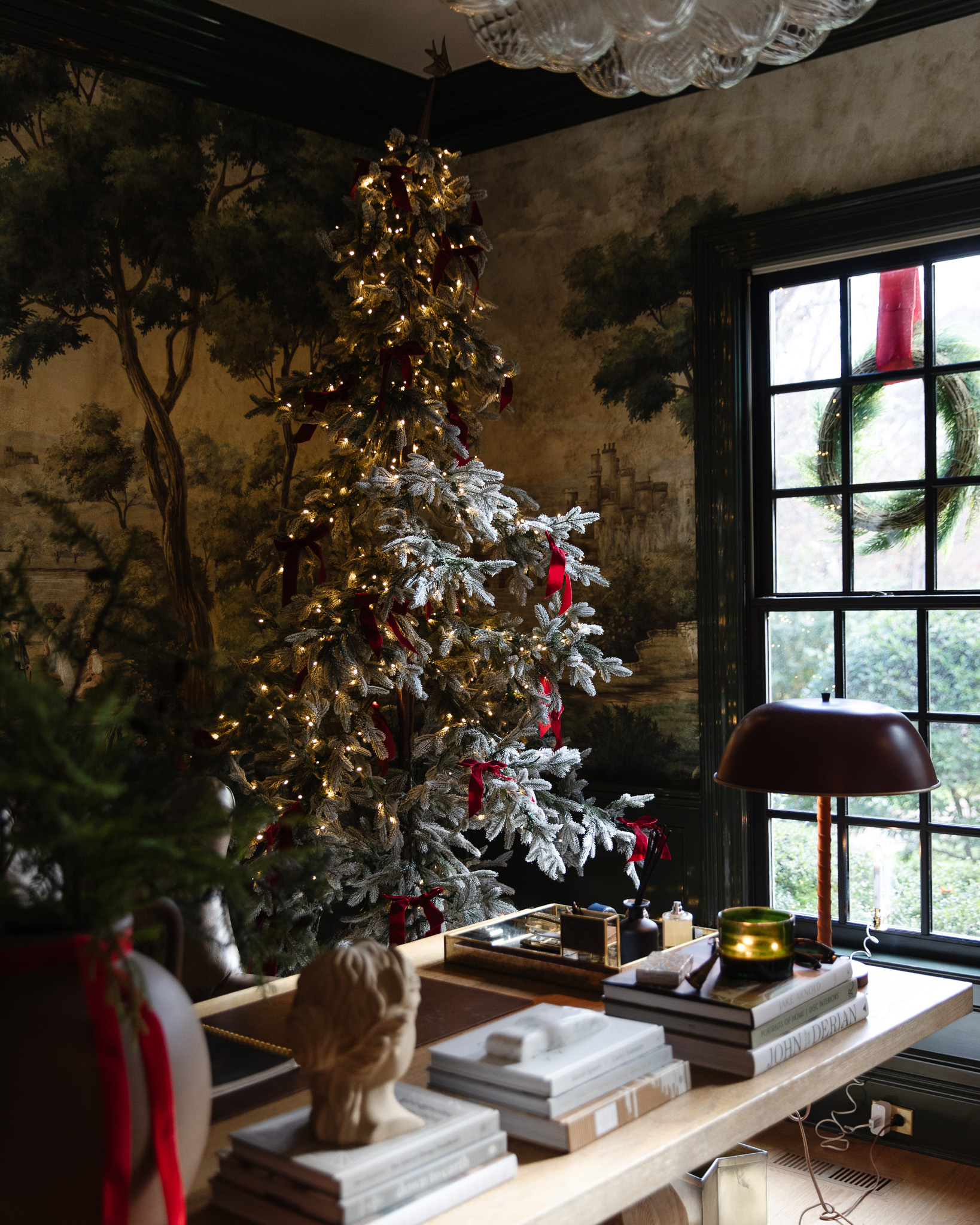 The flocked Christmas tree in the corner of a study with red bows and an outdoor wreath visible from the window