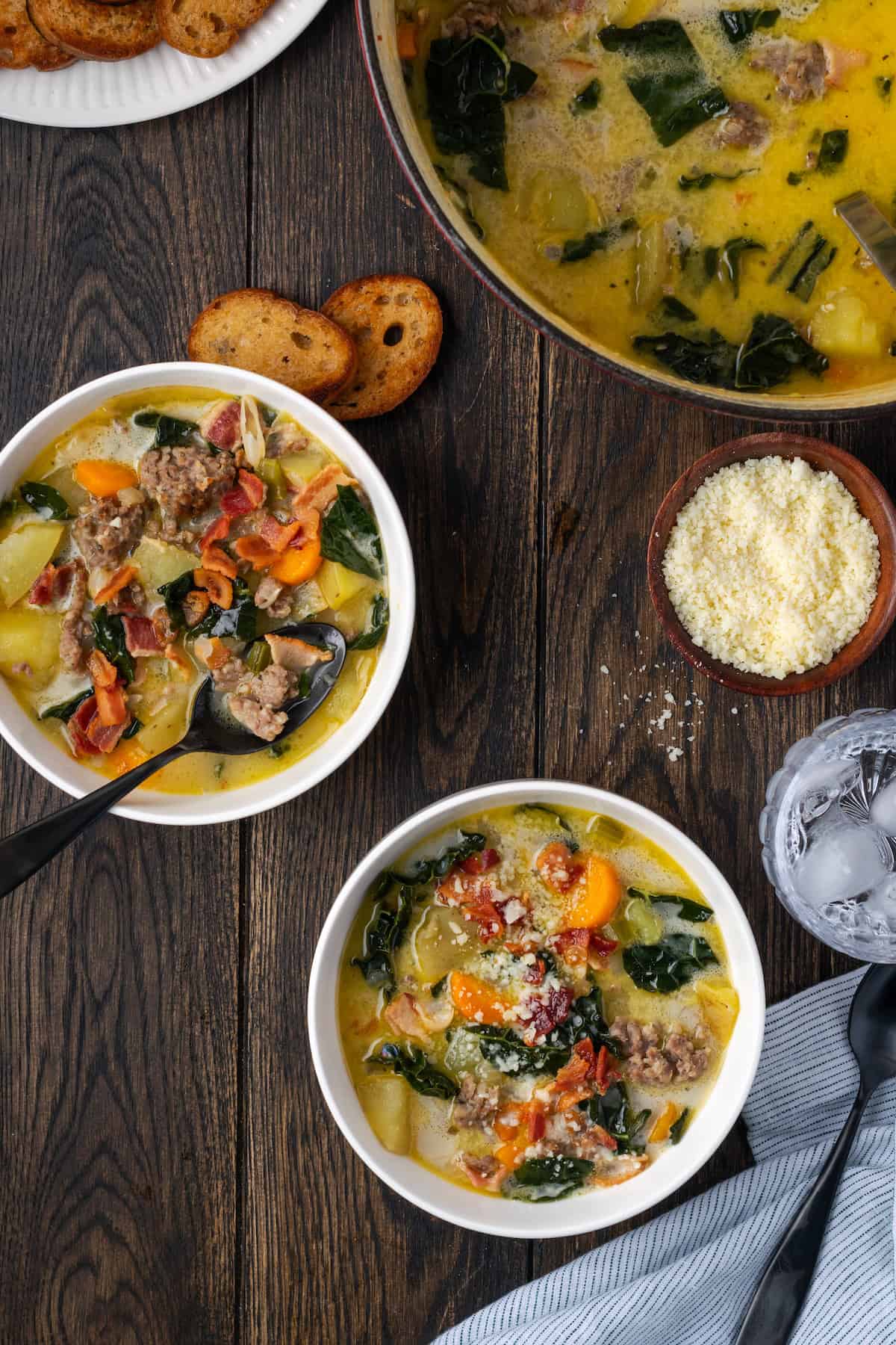 Overhead view of two bowls of Zuppa Toscana next to a small dish of grated parmesan and a large pot of soup.