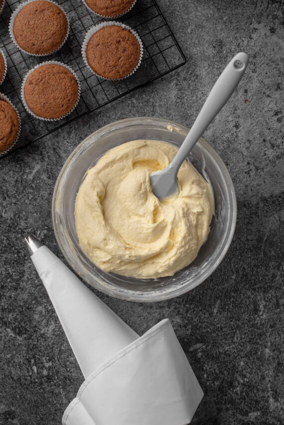 Cream cheese frosting in a bowl with a spatula next to cupcakes on a wire rack.