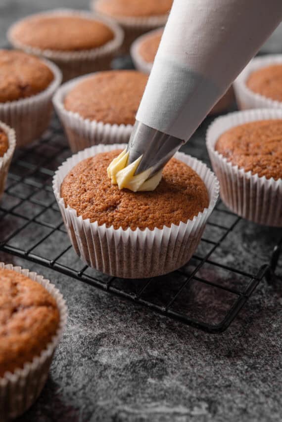 A piping tip piping frosting in the center of a gingerbread cupcake, with more unfrosted cupcakes in the background.