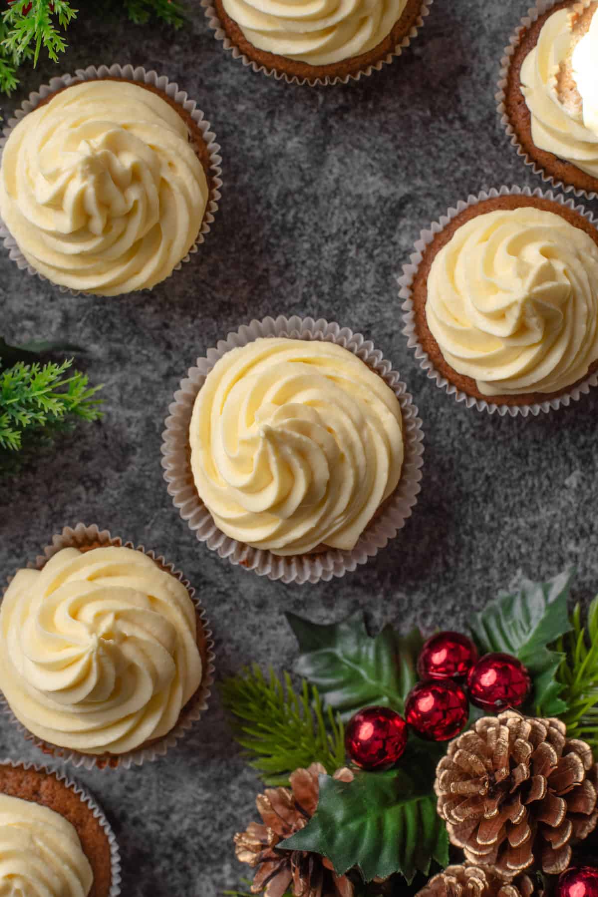 Overhead view of assorted gingerbread cupcakes topped with frosting swirls next to a Christmas table arrangement.