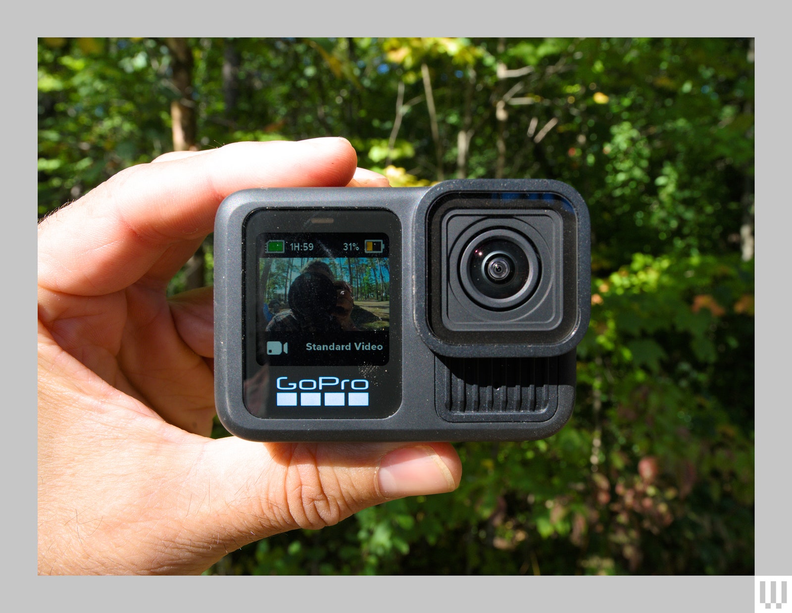 Hand holding small camera showing the screen and lens with bushes in the background