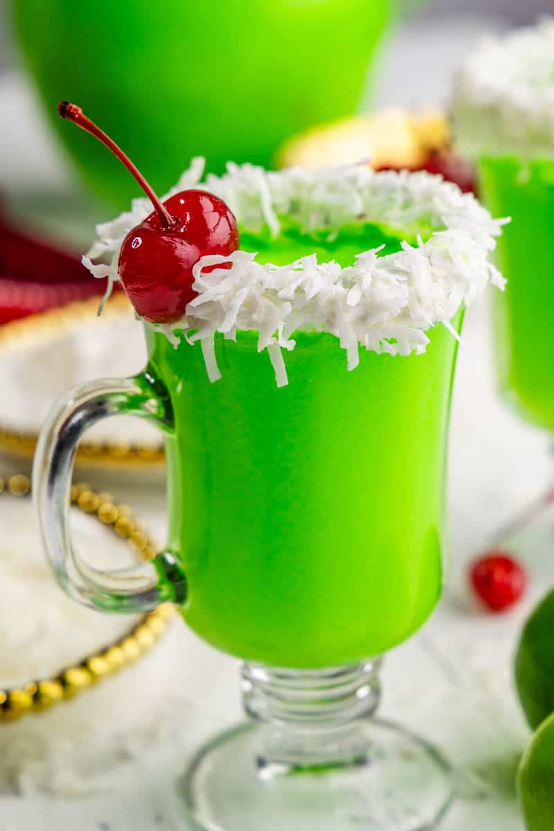 A single glass of green punch in a clear glass mug with a white frosting and coconut rim with a red cherry garnish.