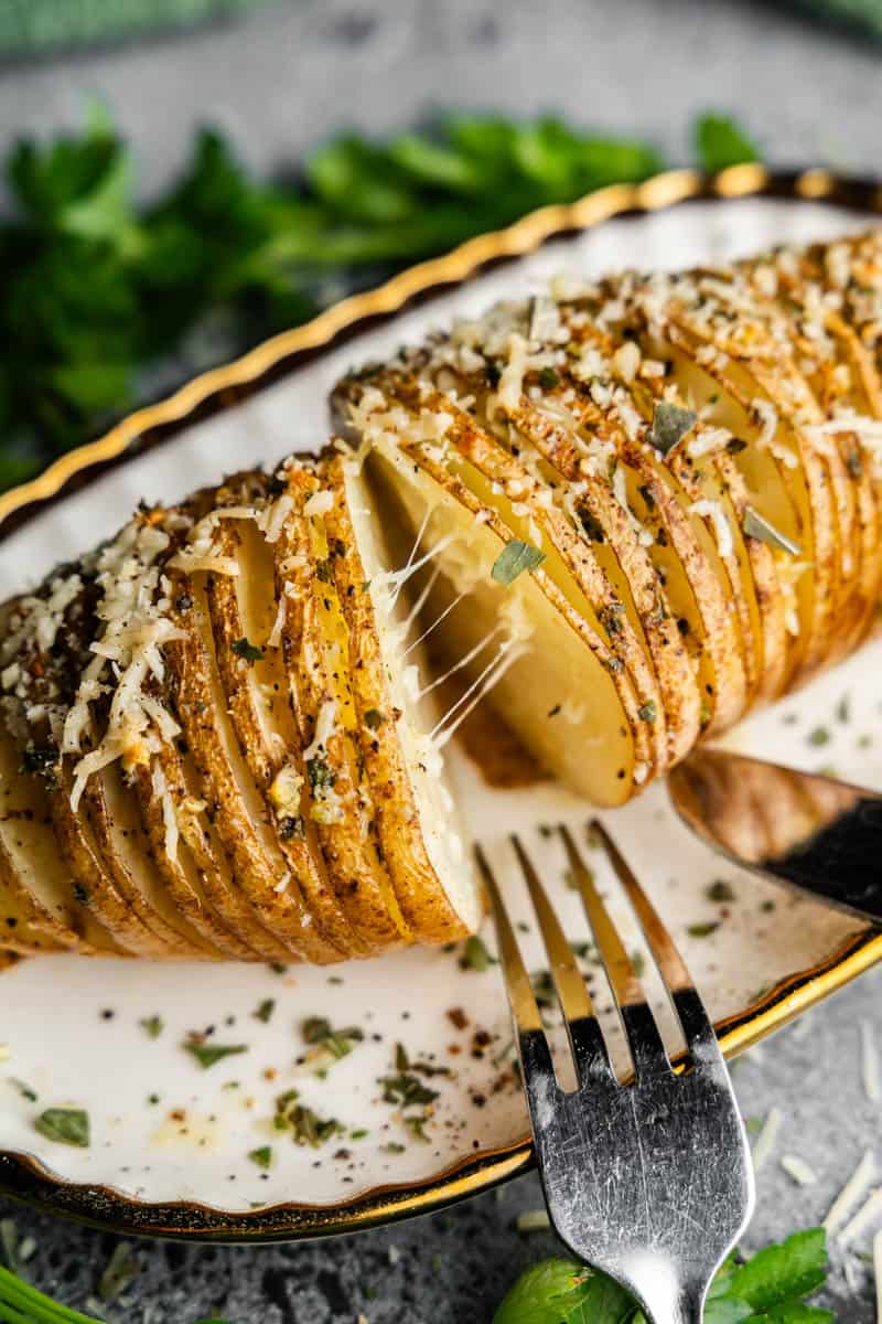 A hasselback potato on a plate with melty cheese, with a fork and knife ready to eat.