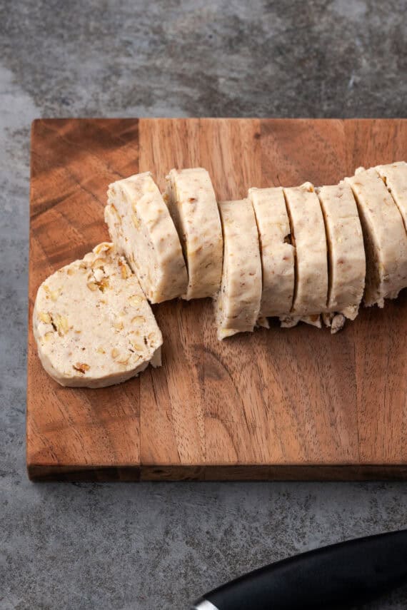 A log of pecan sandies cookie dough sliced into cookies on a wooden cutting board.