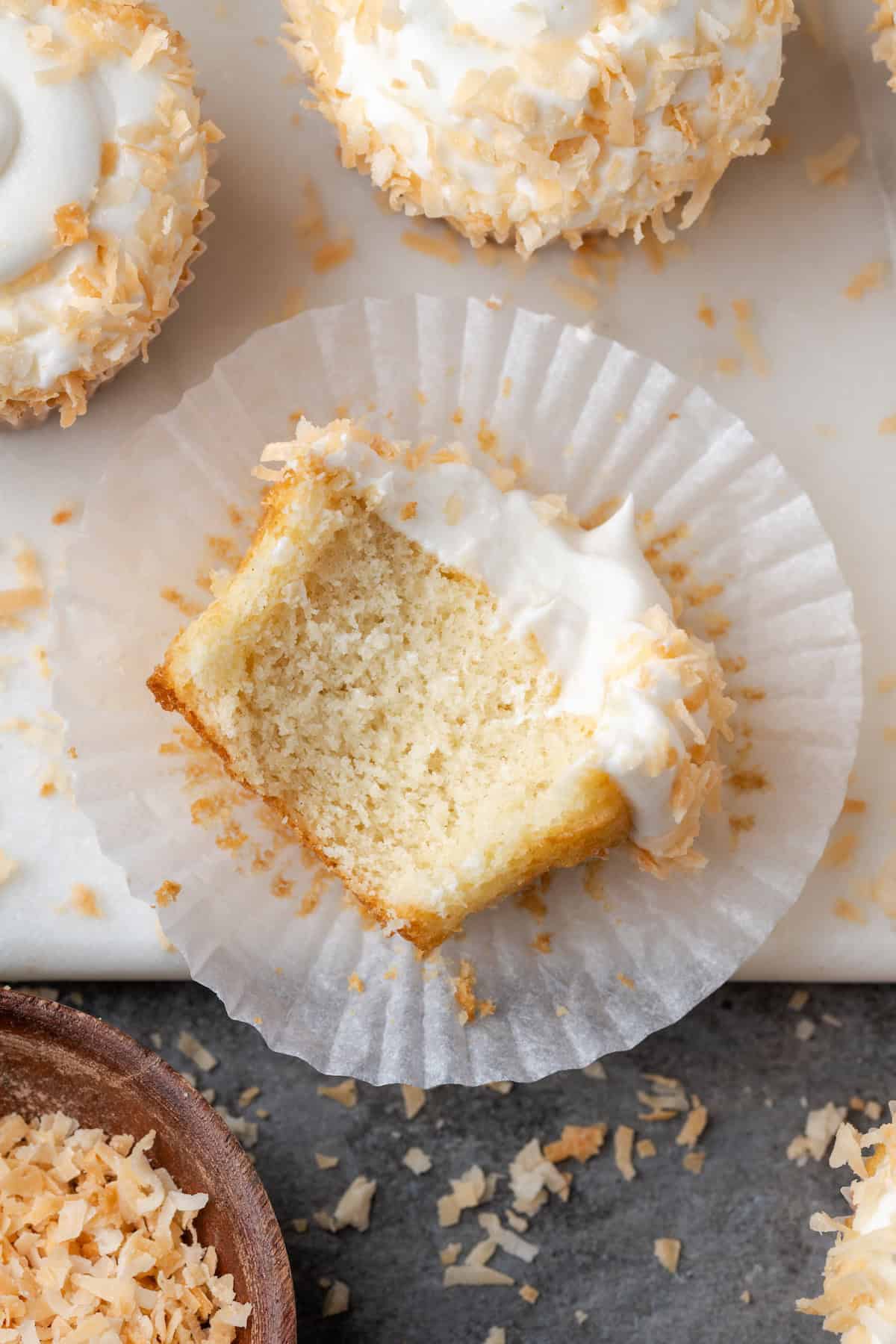 Overhead view of a frosted almond cupcake resting on its side in a cupcake wrapper, with a bite missing.