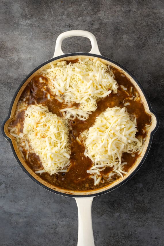 Shredded cheese added overtop chicken breasts in a skillet with onion gravy.