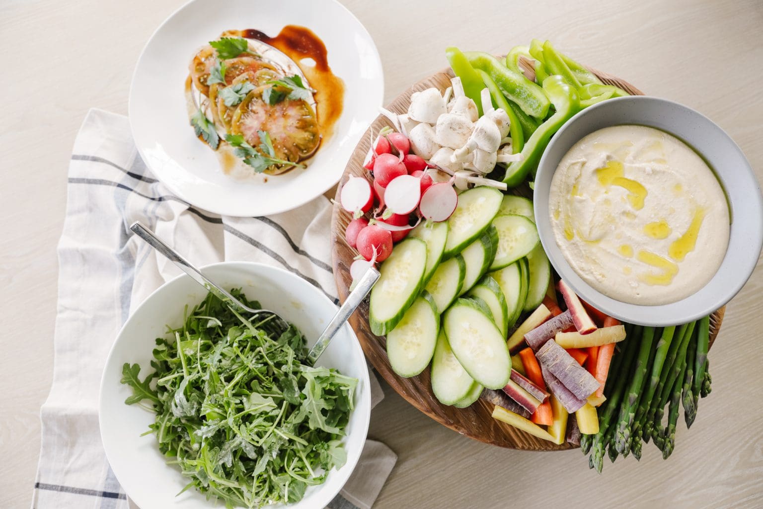 A bowl of hummus next to dippable veggies including asparagus, cucumbers, colorful carroets, radishes, and bell peppers. 