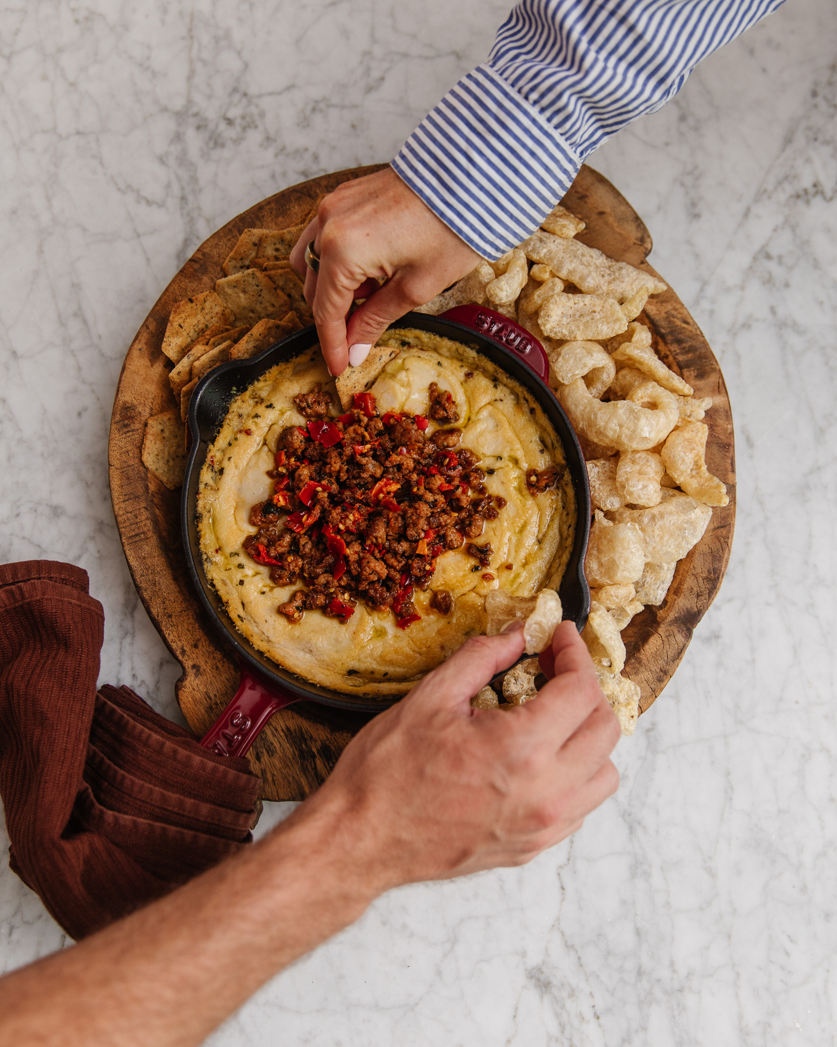 Two hands dipping pork rinds into a spicy bean dip with sausage on top