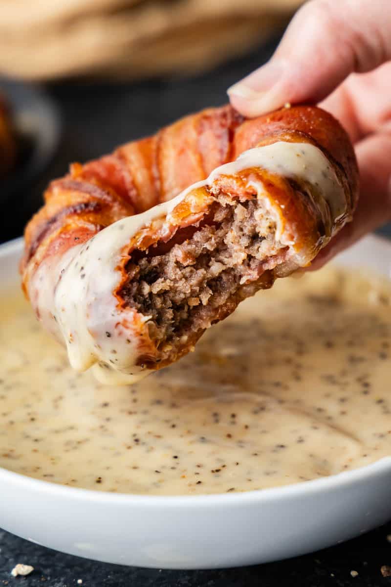 A bacon wrapped sausage filled onion ring being dipped in white sauce with a bite taken out.