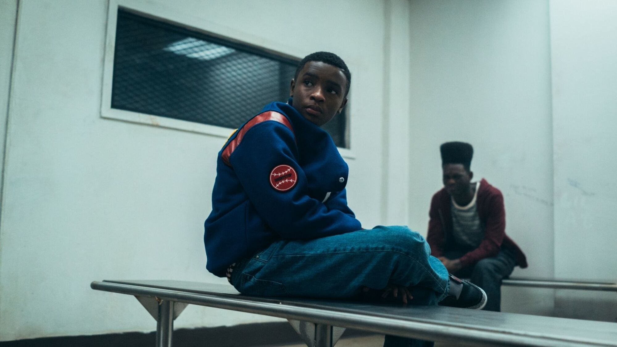 A young Black boy in a baseball jacket sits on a bench with another Black boy in the background, from 