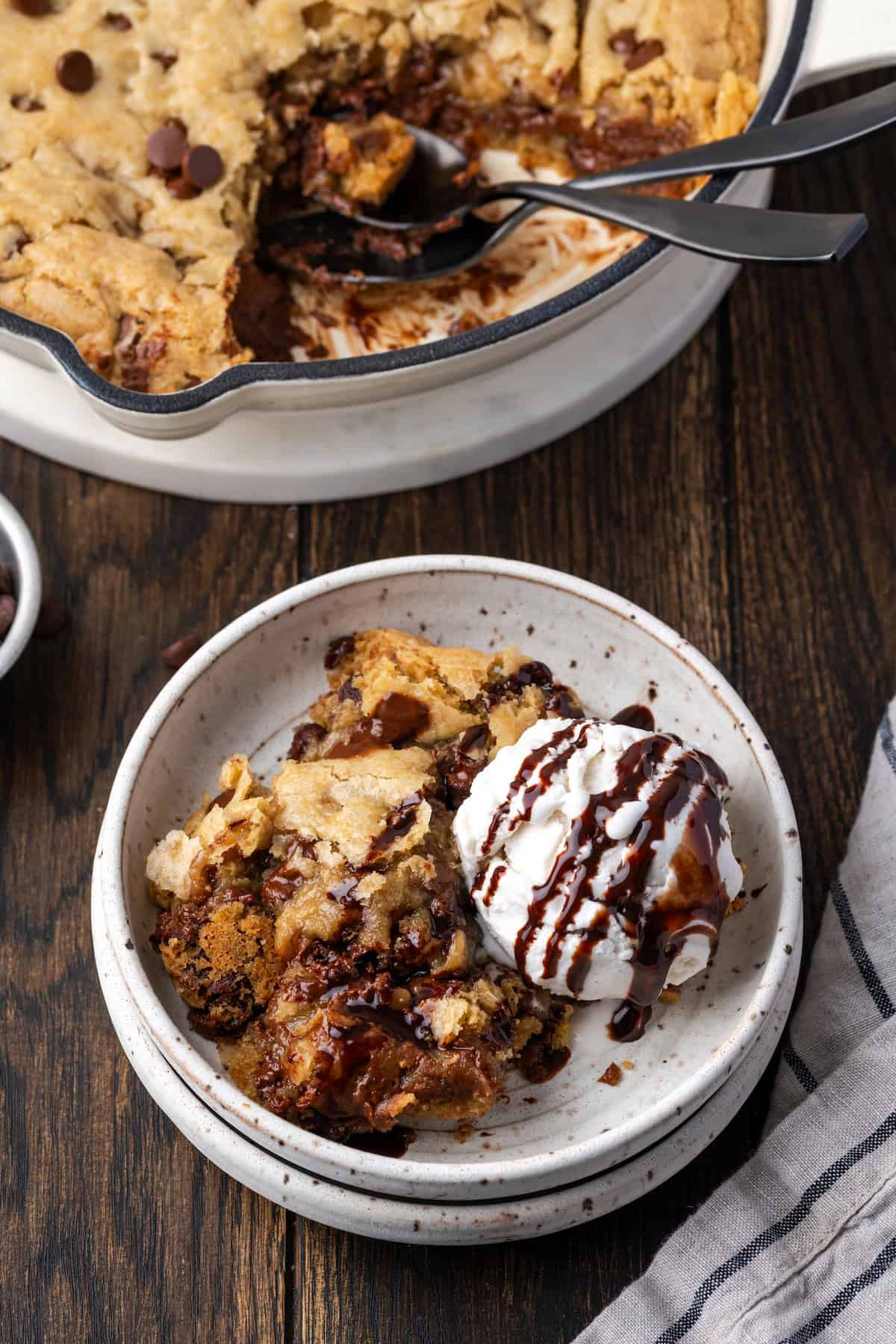 A scoop of chocolate chip skillet cookie in a bowl topped with ice cream and chocolate sauce, next to the rest of the cookie in the skillet.