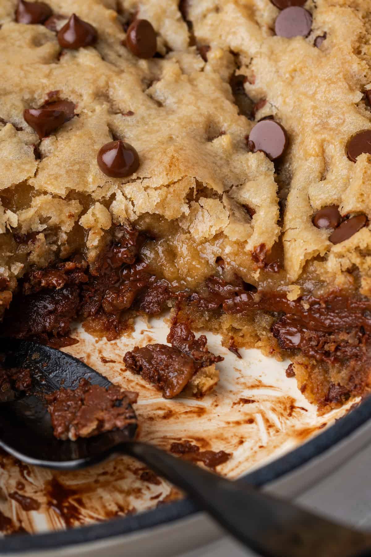 Close up of a skillet chocolate chip cookie with a large serving missing and a spoon resting in the open space.